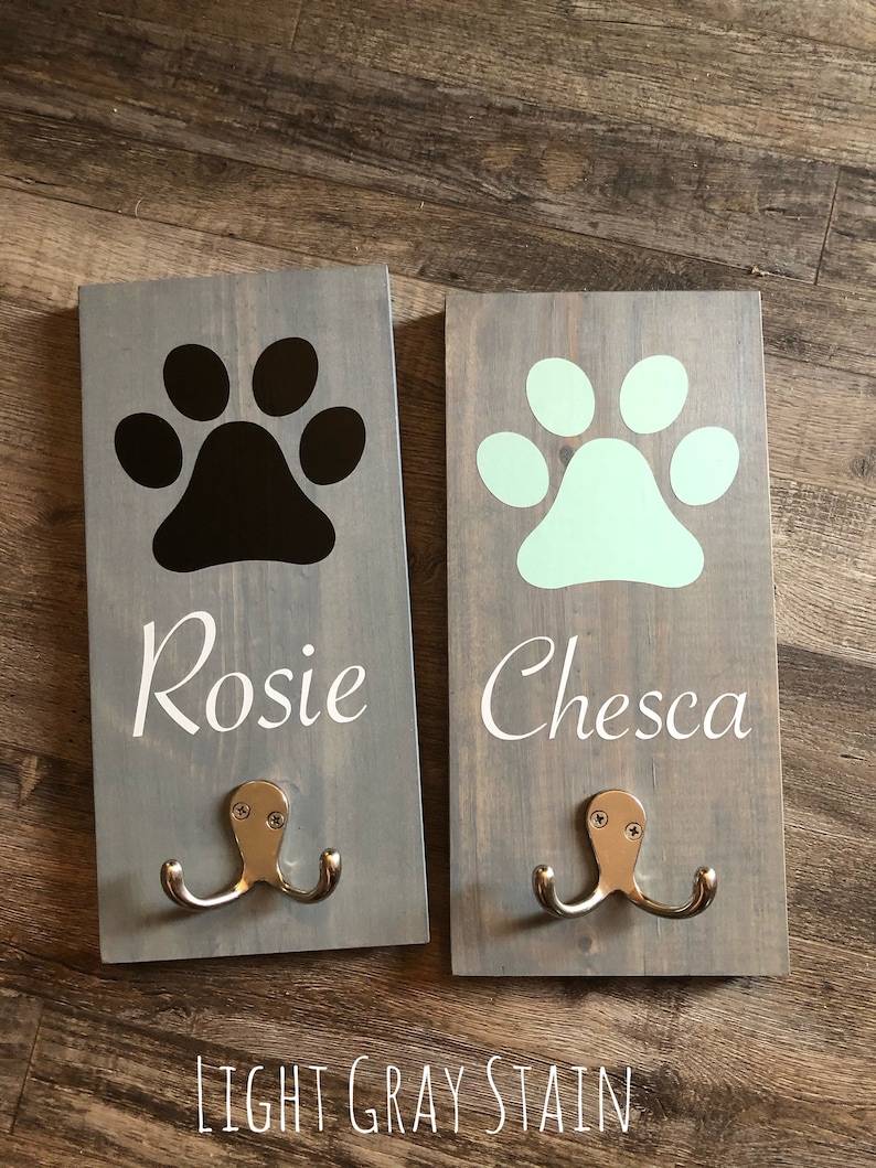 May include: Two wooden dog leash hooks with paw prints and names "Rosie" and "Chesca" in white lettering. The hooks are painted light gray and have silver double hooks.