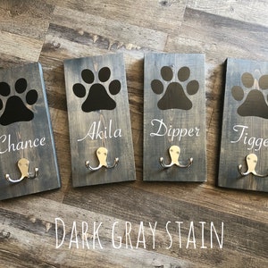 May include: Four wooden dog leash hooks with black paw prints and the names Chance, Akila, Dipper, and Tigger. The hooks are stained dark gray and have two metal hooks each.
