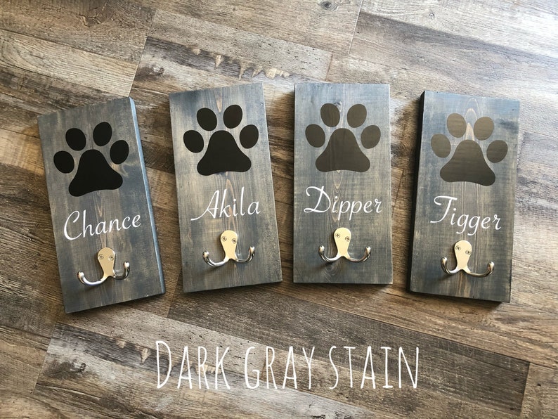 May include: Four wooden dog leash hooks with black paw prints and the names Chance, Akila, Dipper, and Tigger. The hooks are stained dark gray and have two metal hooks each.