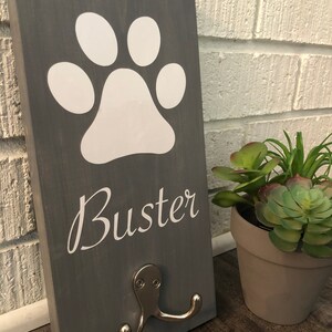 May include: Gray wooden sign with a white paw print and the name "Buster" in white lettering. The sign has a silver double hook.