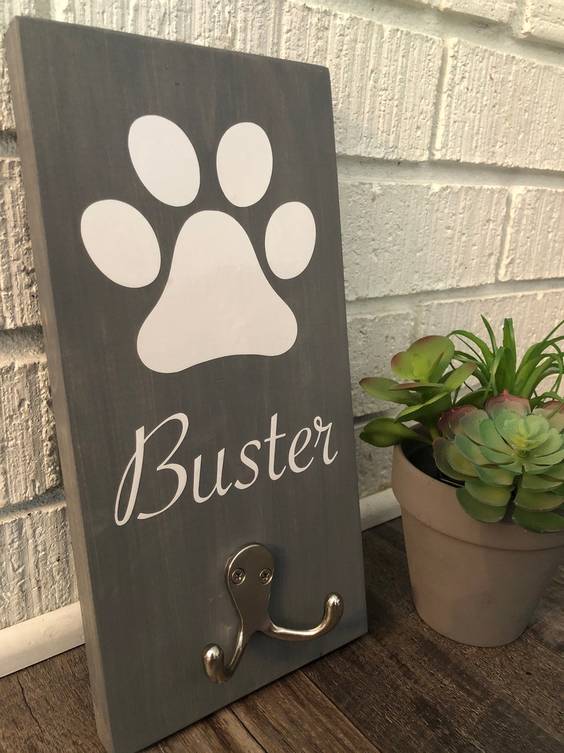 May include: Gray wooden sign with a white paw print and the name "Buster" in white lettering. The sign has a silver double hook.