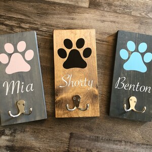 May include: Three wooden dog leash hooks with paw print designs and personalized names. The hooks are painted in gray, brown, and black with white lettering. The names are Mia, Shorty, and Benton.