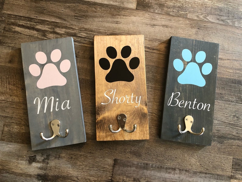 May include: Three wooden dog leash hooks with paw print designs and personalized names. The hooks are painted in gray, brown, and black with white lettering. The names are Mia, Shorty, and Benton.