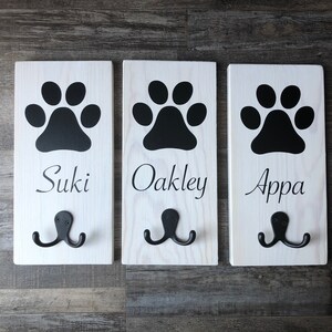May include: Three white wooden dog leash hooks with black paw prints and the names "Suki", "Oakley", and "Appa" written below.