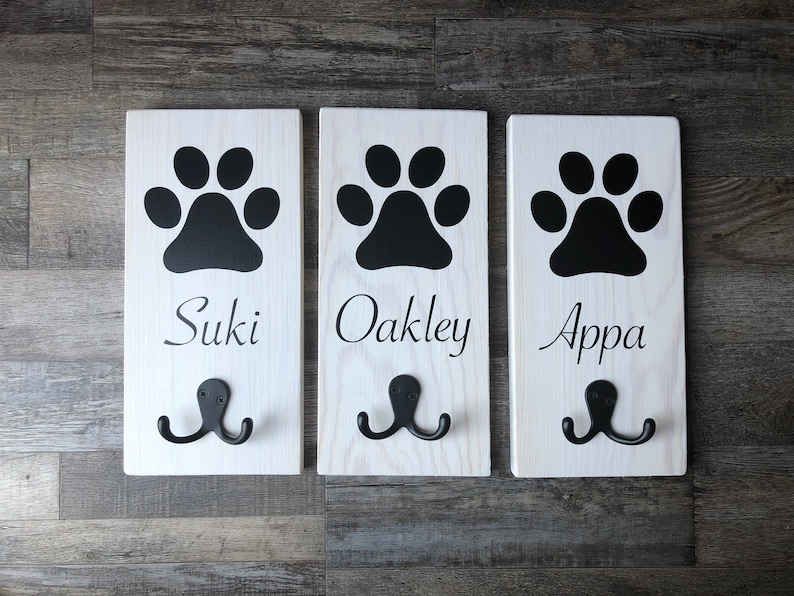 May include: Three white wooden dog leash hooks with black paw prints and the names "Suki", "Oakley", and "Appa" written below.