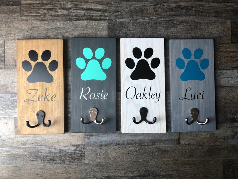 May include: Four wooden dog leash hooks with paw print designs and names "Zeke", "Rosie", "Oakley", and "Luci" painted on them. The hooks are mounted on a wall.