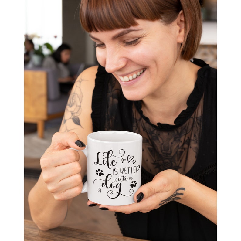Puede incluir: Taza de caf&eacute; de cer&aacute;mica blanca con un dise&ntilde;o negro que dice "La vida es mejor con un perro" con huellas de patas y corazones.