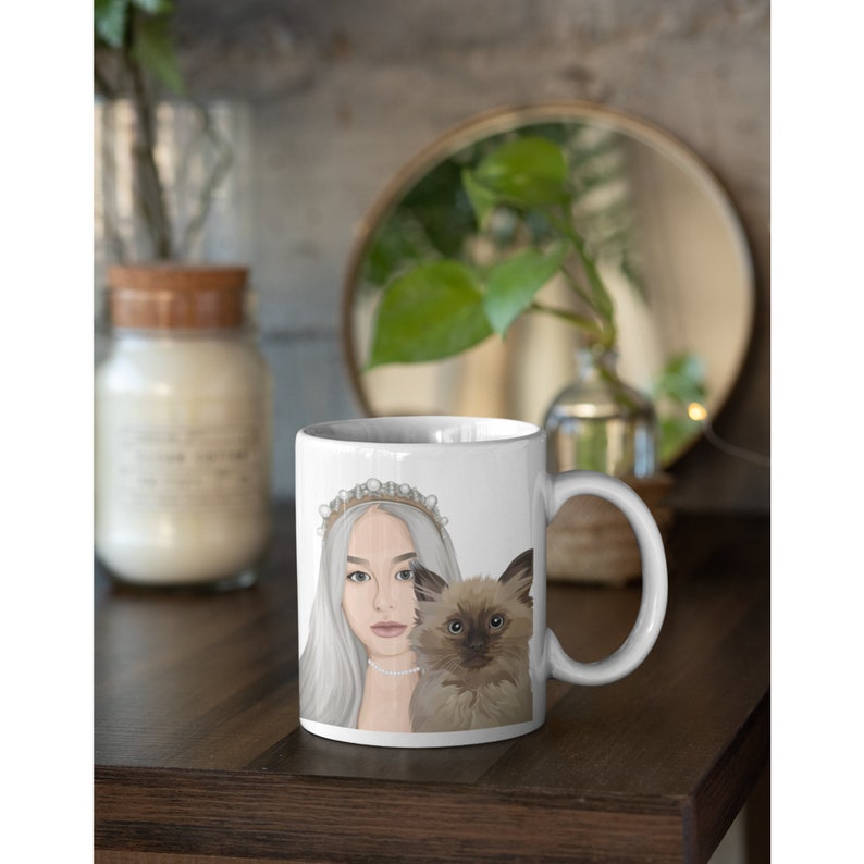 Puede incluir: Taza de cer&aacute;mica blanca con una ilustraci&oacute;n colorida de una mujer con una corona y un gato. La mujer tiene el pelo largo y rubio y lleva un vestido blanco. El gato es marr&oacute;n y blanco y est&aacute; sentado junto a la mujer.