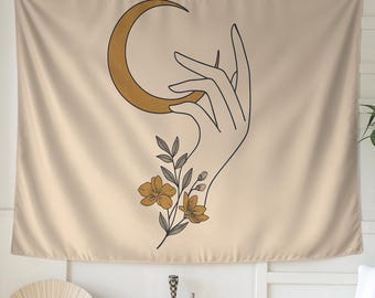 May include: Beige tapestry featuring a hand holding a crescent moon. The hand is outlined in black, with a gold-colored moon. Below the hand is a floral arrangement with yellow flowers and gray leaves. The tapestry is a decorative wall hanging.
