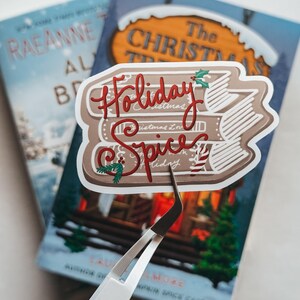 May include: A sticker with the words "Holiday Spice" in red script, layered over a stack of books. The sticker is held by tweezers over two books with blue and white covers. The books have titles like "The Christmas Tree" and "RaeAnne" on them.