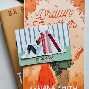 May include: An image of three books, one with the title "Drawn Together" and another with the author's name "Juliana Smith". A bookmark with a cartoon illustration of a person reading and the word "BOOKMARK" is placed on top of the books.