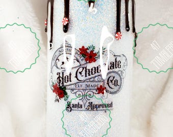 Puede incluir: Un vaso blanco con purpurina con un diseño de goteo de chocolate y caramelos de menta. El vaso presenta el texto "Hot Chocolate Co. Elf Made Santa Approved" con acentos florales rojos. Un artículo de bebida festivo.