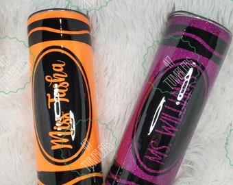 May include: Two glittery tumblers designed to resemble crayons. One is orange with black stripes and the name "Miss Trisha" in a cursive font. The other is purple with black stripes and the text "MS. WILL" in a similar font.