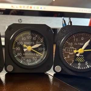 May include: Three black desk organisers shaped like aircraft instrument panels. The left panel has a compass with a red aeroplane graphic. The middle panel has an airspeed indicator with the text "AIRSPEED" and "KNOTS". The right panel has an altimeter with the text "ALTIME" and "1013".