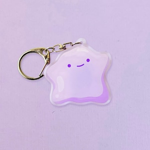 May include: A light purple, star-shaped keychain with a smiling face. The keychain has a gold-colored keyring and clasp. The design features a lighter purple outline and a darker purple face. The background is a solid light purple.