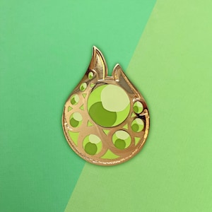 Könnte beinhalten: Goldfarbene Emaille-Anstecknadel in Tropfenform mit einem zentralen grünen Edelstein und kleineren grünen Akzenten. Die Anstecknadel hat ein detailliertes Design mit ausgeschnittenen Formen und ein glänzendes Finish, vor grünem Hintergrund.