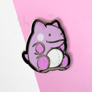 May include: A lavender enamel pin featuring a cartoon-style creature with a white belly and a silver outline. The pin has a white circle on its head and two pink circles on its body. The background is pink and white.