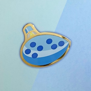 Peut inclure: Épinglette ocarina bleu clair avec des détails de points bleu foncé et bordure dorée. Le design stylisé comprend une bordure dorée et un petit emblème triangulaire jaune. Le fond est un dégradé de bleu clair et moyen.
