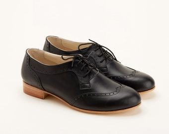 May include: A pair of black leather oxford shoes featuring classic brogue detailing with perforated patterns along the toe cap and sides. The shoes have thin black laces, a low stacked wooden heel, and a light-colored interior lining.