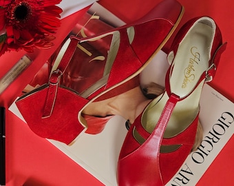 May include: A pair of red heeled shoes with a T-strap design. The shoes are made of leather and suede and have a buckle closure. The shoes are sitting on a magazine with the text "Giorgio Armani" printed on the cover.
