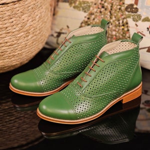Puede incluir: Un par de botines de cuero verde con un diseño perforado y cordones marrones. Los botines tienen una punta redonda y un tacón bajo.