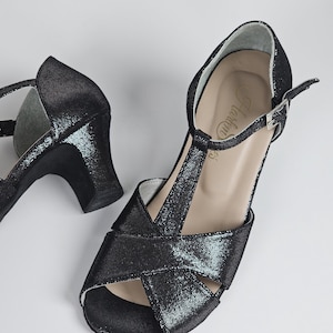 Könnte beinhalten: Schwarze Glitzer-Sandalen mit offenem Zehenbereich, T-Riemen und Knöchelriemen. Die Absätze sind mittelhoch. Die Sandalen haben ein strukturiertes Aussehen und einen Schnallenverschluss. Die Schuhe sind für formelle Anlässe bestimmt.