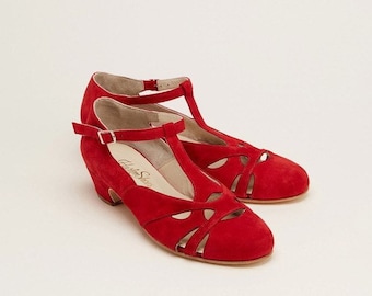 May include: Red suede heeled shoes with a T-strap and cut-out design. The shoes have a low block heel and a buckle closure.
