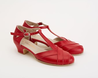 May include: A pair of red leather heeled shoes with a t-strap design. The shoes have a low block heel and a pointed toe.