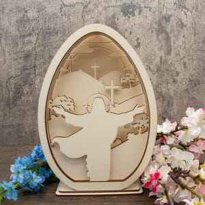 May include: A wooden Easter egg-shaped ornament with a cutout design of Jesus Christ with outstretched arms, standing on a hill with three crosses behind him. The ornament is light brown and has a white background.