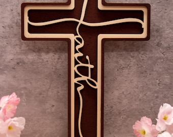 May include: A wooden cross with the word "faith" carved into it. The cross is brown with a light brown border. The cross is standing on a wooden surface with pink flowers in the foreground.