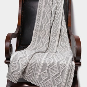 May include: A gray cable knit throw blanket with a diamond pattern draped over a wooden armchair with a black leather seat.