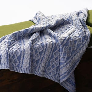May include: A blue and white cable knit blanket draped over a green cushion. The blanket features a detailed pattern of interwoven cables and diamond shapes. The cozy throw is made of soft material.