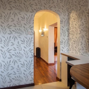 Puede incluir: Interior de cocina con armarios blancos, horno empotrado y paredes con papel pintado con estampado de hojas grises. Una mesa de madera con un cuenco blanco con fruta está en primer plano. El ambiente es cálido y acogedor.
