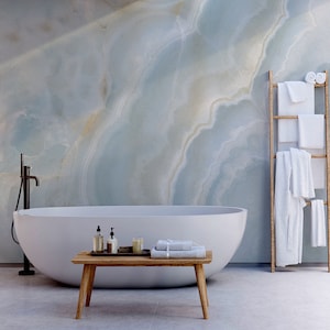 May include: A white bathtub with a wooden bench in front of it. The bathroom has a marble wall and a wooden ladder with white towels hanging on it. A glass door leads to an outdoor shower with a wooden wall and a green plant.