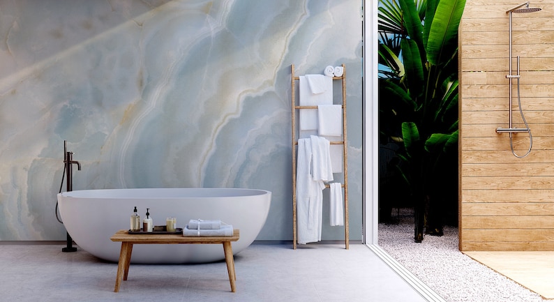 May include: A white bathtub with a wooden bench in front of it. The bathroom has a marble wall and a wooden ladder with white towels hanging on it. A glass door leads to an outdoor shower with a wooden wall and a green plant.