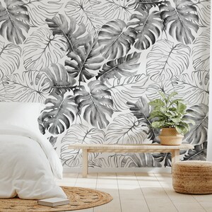 May include: A bedroom interior with a white bed and a white throw blanket. The wall is covered in a black and white tropical leaf wallpaper. A wooden bench and a woven basket are in the foreground.