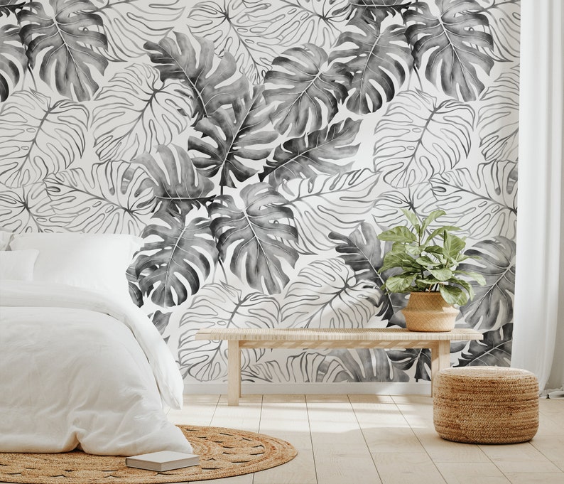 May include: A bedroom interior with a white bed and a white throw blanket. The wall is covered in a black and white tropical leaf wallpaper. A wooden bench and a woven basket are in the foreground.