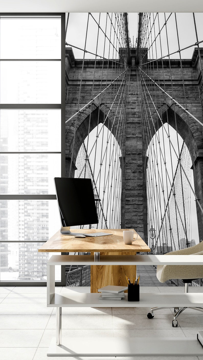 May include: A black and white photo of the Brooklyn Bridge in New York City. The photo is used as a wall mural in a modern office space with a white desk, a computer, and a chair.