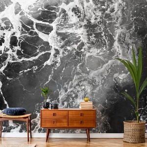 May include: A black and white abstract wallpaper resembling ocean waves. A wooden mid-century modern dresser, a woven chair with a blue cushion, and potted plants are in front of the wallpaper. The room has a hardwood floor.
