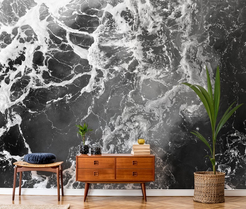 May include: A black and white abstract wallpaper resembling ocean waves. A wooden mid-century modern dresser, a woven chair with a blue cushion, and potted plants are in front of the wallpaper. The room has a hardwood floor.