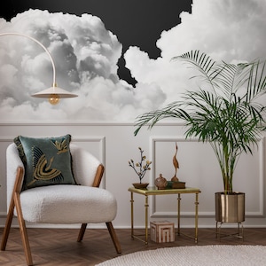 Puede incluir: Un sillón blanco con un cojín estampado en azul verdoso y dorado se encuentra en una sala de estar con un papel tapiz de nubes en blanco y negro, una palmera en maceta y una mesa auxiliar dorada con objetos decorativos.