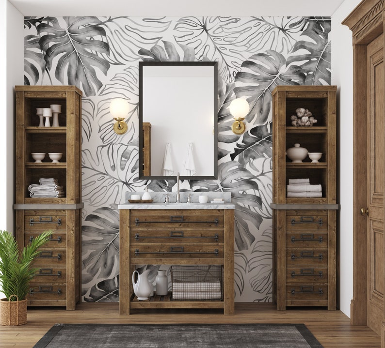May include: A bathroom vanity with a white countertop and wooden cabinets. The vanity has a large mirror above it and two sconces on either side. The walls are covered in a black and white tropical leaf wallpaper.