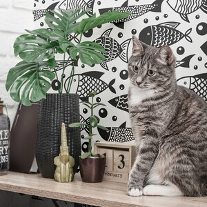 May include: A gray tabby cat with white markings sits on a wooden shelf in front of a black and white wallpaper with a fish pattern. The wallpaper has the text "Home is where our story begins". There are also two potted plants and a gold elephant figurine on the shelf.