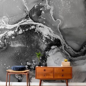 May include: A black and white abstract marble wallpaper with a silver metallic sheen. A wooden dresser with four drawers and a stool with a blue velvet cushion are in front of the wallpaper.
