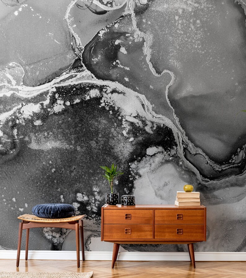 May include: A black and white abstract marble wallpaper with a silver metallic sheen. A wooden dresser with four drawers and a stool with a blue velvet cushion are in front of the wallpaper.
