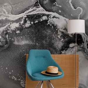 May include: A teal blue chair with a straw hat on the seat. The chair has a silver metal base and is in front of a wall with a black and white abstract design. A white lamp with a silver base is in the background.