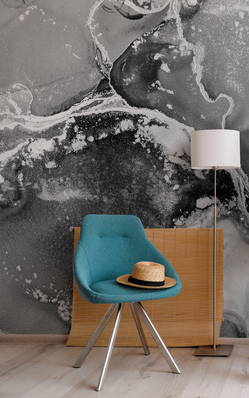 May include: A teal blue chair with a straw hat on the seat. The chair has a silver metal base and is in front of a wall with a black and white abstract design. A white lamp with a silver base is in the background.