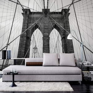 May include: A black and white photograph of the Brooklyn Bridge in New York City. The bridge is shown from a low angle, with the cables and towers visible. The photo is used as a wall mural behind a gray sofa with brown throw pillows.