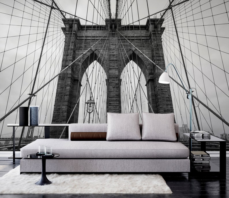 May include: A black and white photograph of the Brooklyn Bridge in New York City. The bridge is shown from a low angle, with the cables and towers visible. The photo is used as a wall mural behind a gray sofa with brown throw pillows.