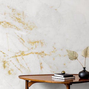 May include: A white marble background with gold accents. A wooden table with a black vase with dried palm leaves and a stack of books.